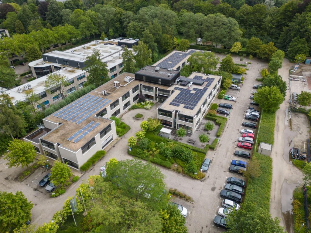Luchtfoto van het gebouw aan de Burgemeester Verderlaan met zonnepanelen op het dak en een naastgelegen parkeerterrein.
